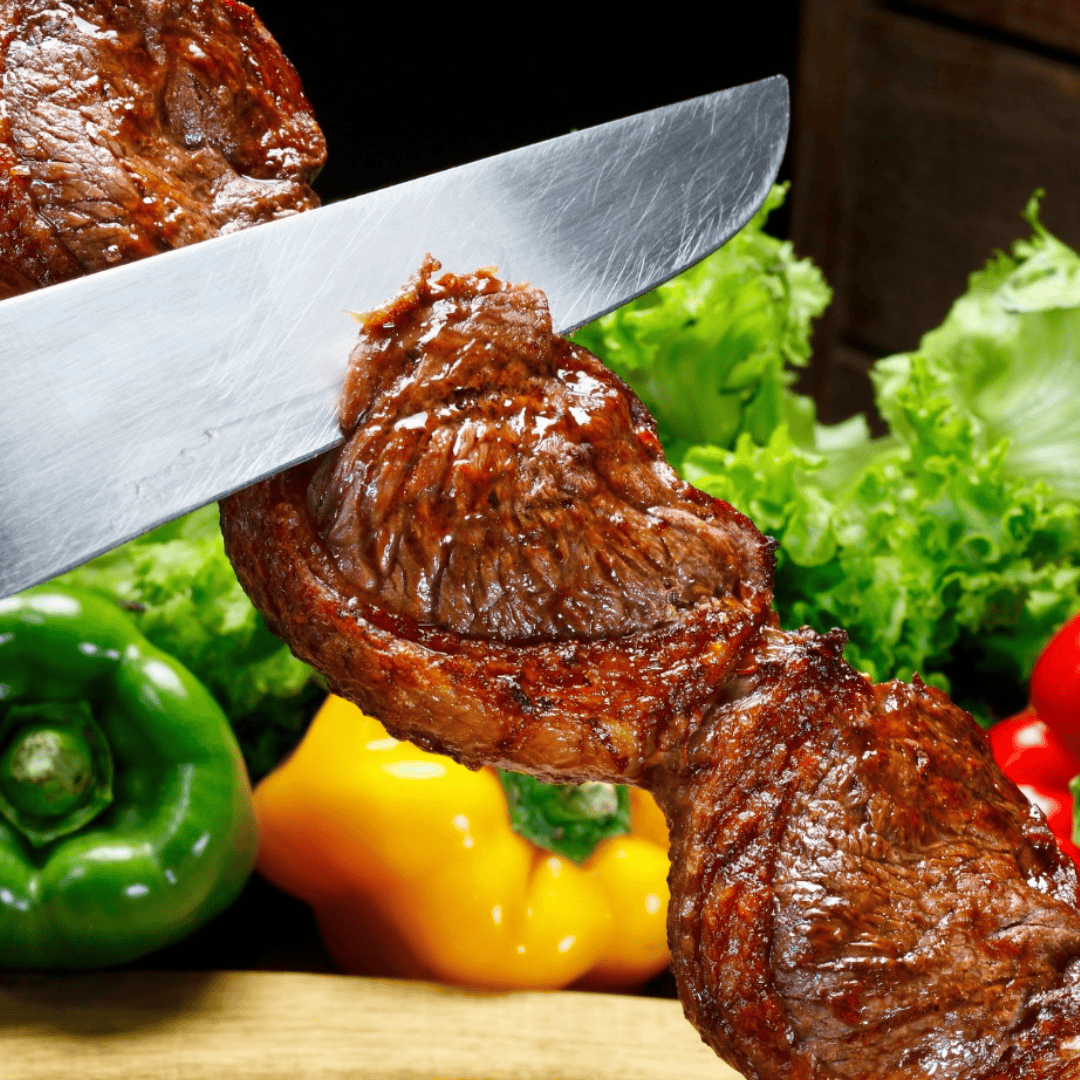 A knife slices grilled meat on a skewer, with green and yellow bell peppers, lettuce, and red tomatoes in the background.