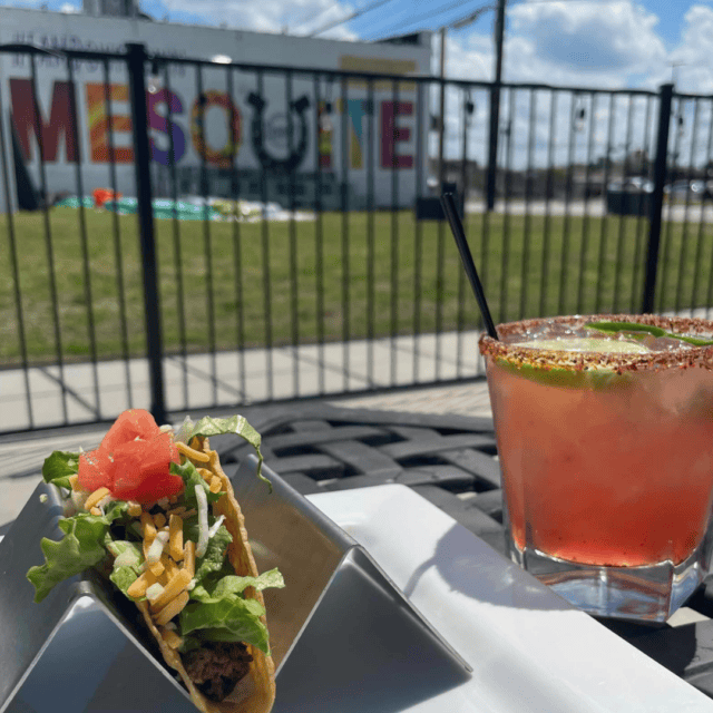 A taco with lettuce, cheese, and tomato sits in a metal holder next to a pink cocktail on an outdoor table, with a MESQUITE mural in the background.