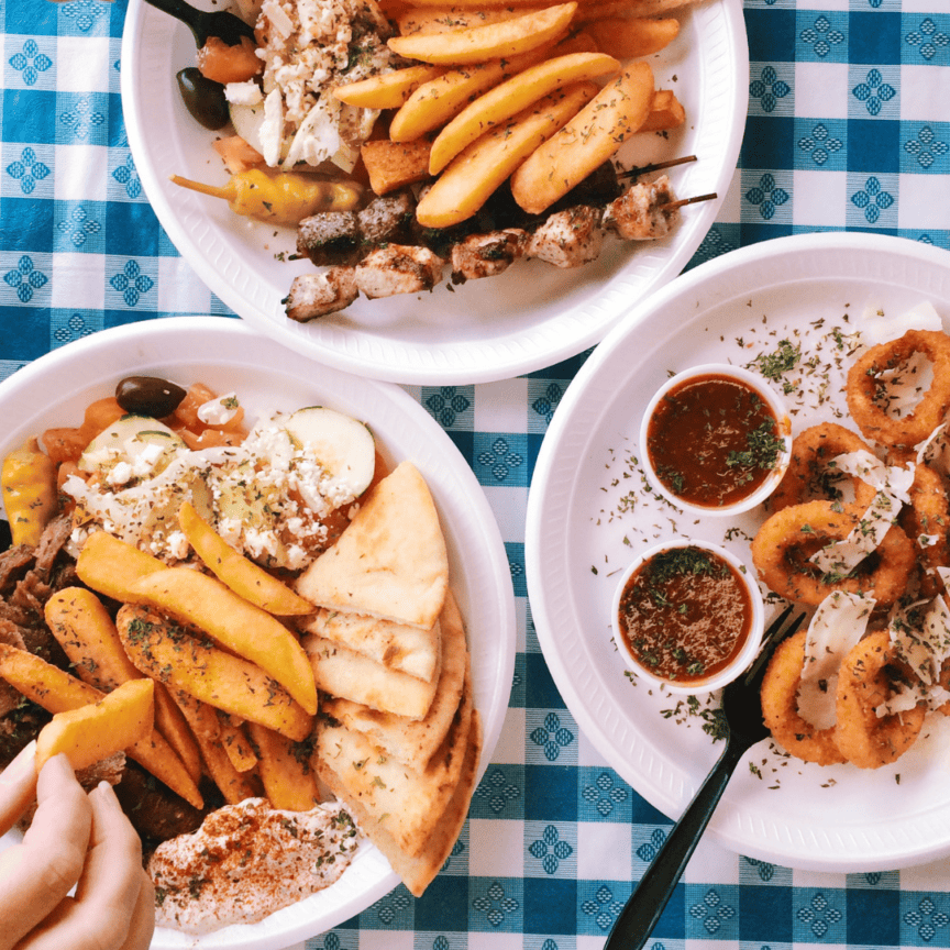 Three plates on a blue and white tablecloth with fries, pita, grilled meat skewers, salad, calamari rings, and dipping sauces. A hand reaches for food on one plate.