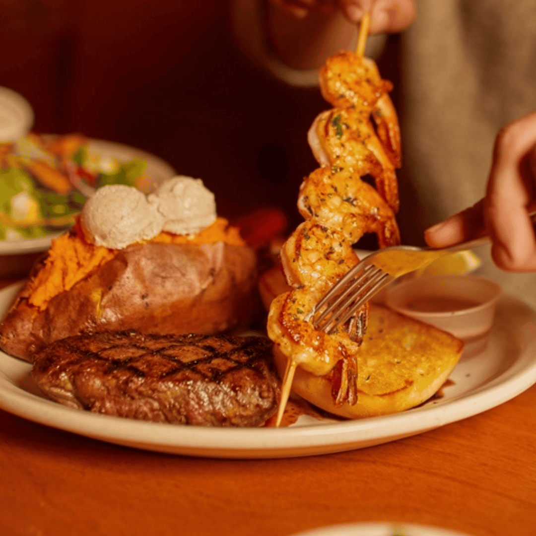 A plate with grilled steak, shrimp skewer, baked sweet potato with butter, garlic bread, and a side salad in the background. A hand holds a fork and lifts the shrimp skewer.