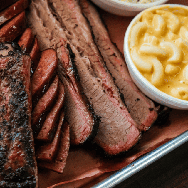 Sliced brisket and sausage served on a tray with bowls of macaroni and cheese and coleslaw.