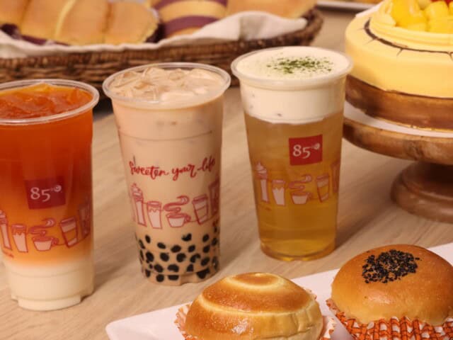 Four drinks and two buns are displayed on a table, with more bread and a fruit-topped cake in the background. The drinks include bubble tea, iced tea, and a milk foam tea.