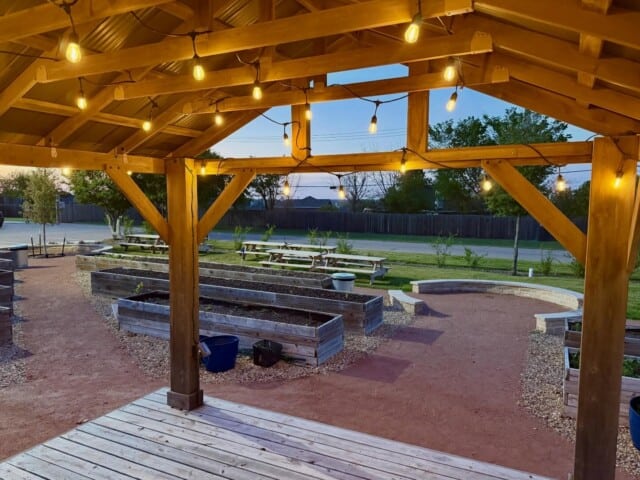 Covered wooden patio with string lights overlooking raised garden beds and gravel seating area in an outdoor setting at dusk.