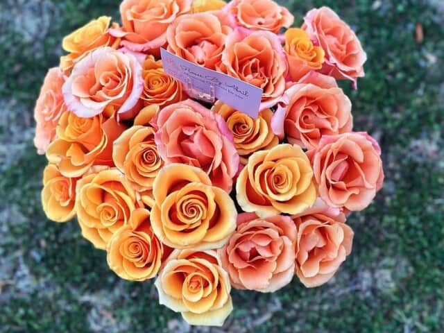 A bouquet of orange and yellow roses with a small pink card in the center, placed on grass outdoors.