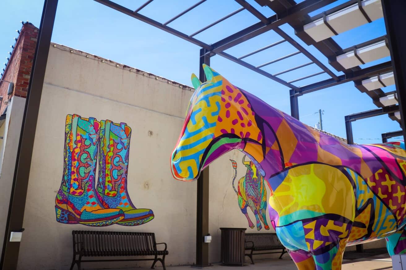 A brightly painted horse sculpture stands under a glass canopy beside a mural of colorful cowboy boots and a lassoing figure on a wall.