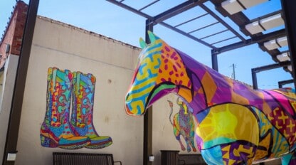A brightly painted horse sculpture stands under a glass canopy beside a mural of colorful cowboy boots and a lassoing figure on a wall.