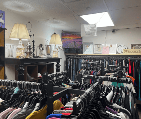 Clothing racks filled with assorted clothes in a thrift store, with lamps, framed art, and decor items displayed on shelves above.