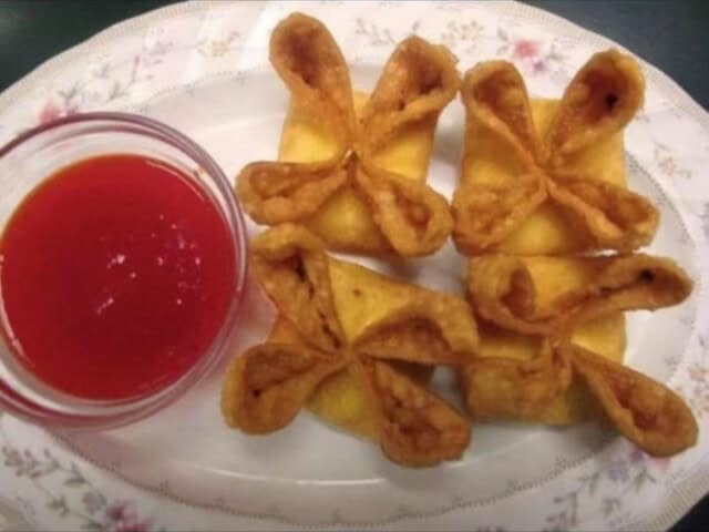 Four fried wontons arranged in a cross shape on a floral plate, with a small glass bowl of red dipping sauce on the side.