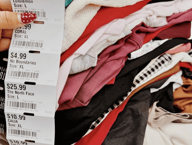 A hand holds up a stack of price tags next to a pile of folded clothing, showing various brands, prices, and sizes.