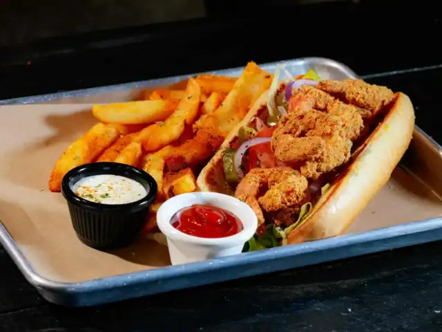 A fried shrimp po’ boy sandwich with lettuce and tomato, served with seasoned fries, ketchup, and a side of ranch dipping sauce on a metal tray.