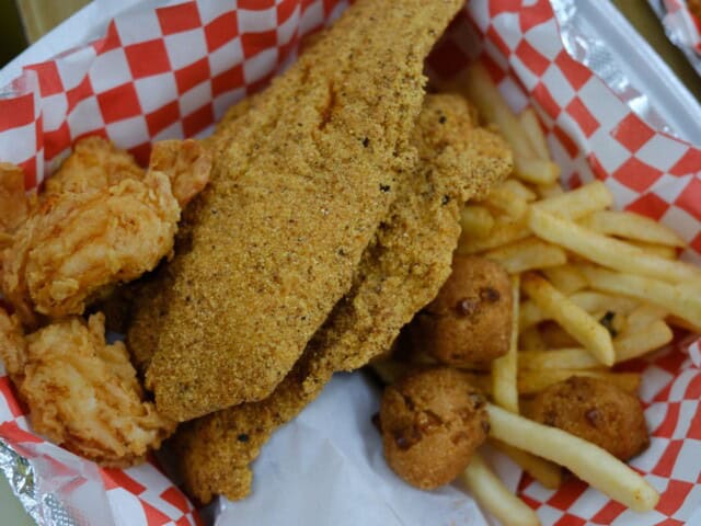 A basket with fried catfish fillets, two pieces of fried chicken, French fries, and hush puppies on red and white checkered paper.