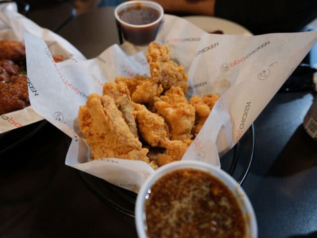 A basket of fried chicken pieces on branded paper with two cups of dipping sauce on a dark table.