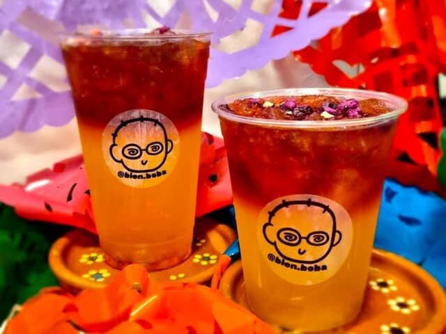 Two plastic cups with layered iced drinks on wooden coasters, surrounded by colorful paper decorations. Each cup features a cartoon face logo and the text “@elon.boba”.